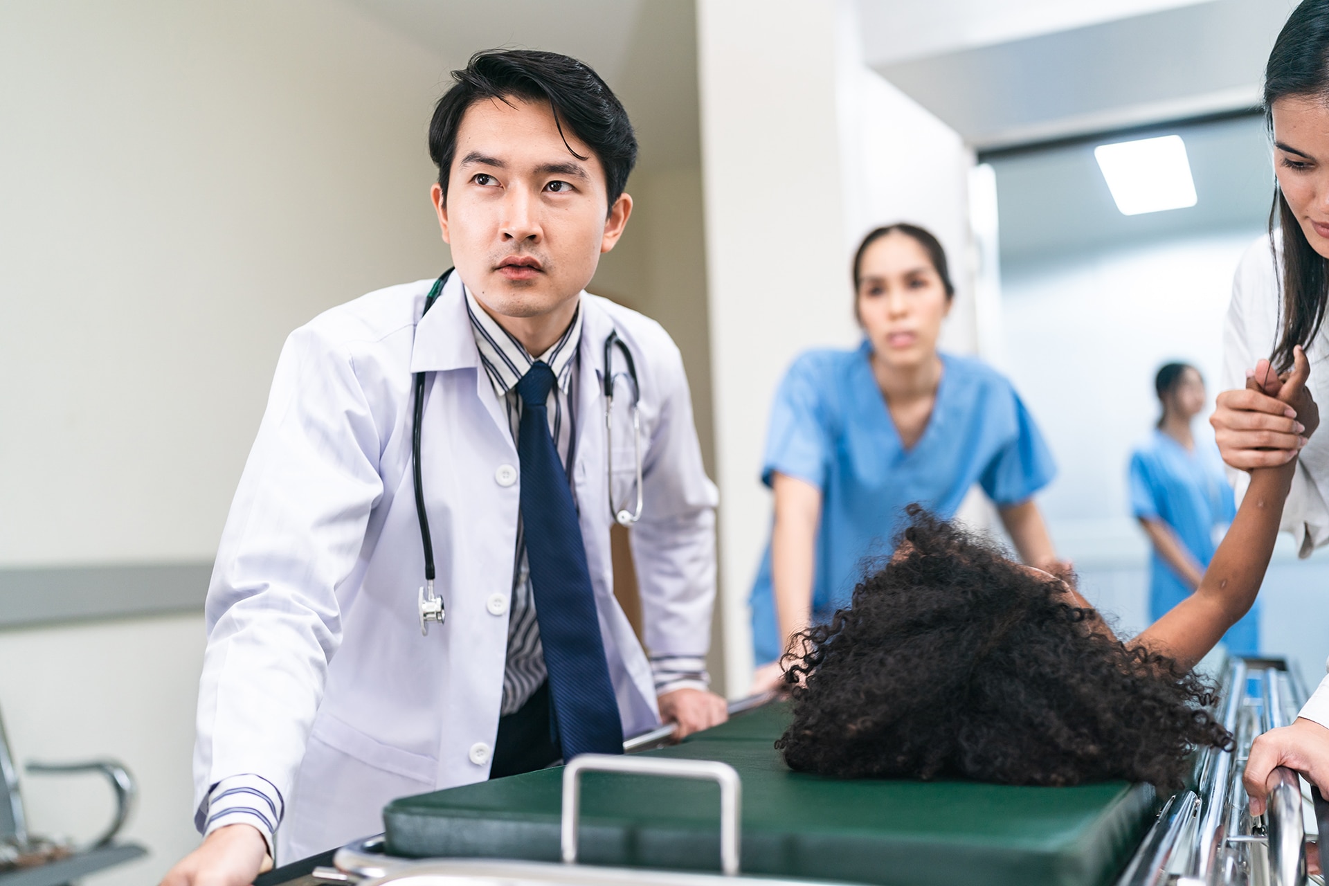 Portrait of young handsome Asian male doctor rushing in hospital during emergency with patient lying on bed with mother holding hand and nurse assisting in corridor passage